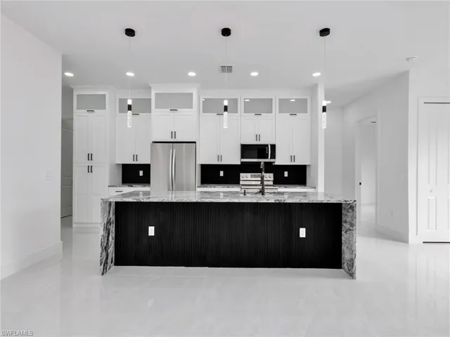 Kitchen with light stone counters, white cabinets, a large island with sink, stainless steel appliances, and glass insert cabinets