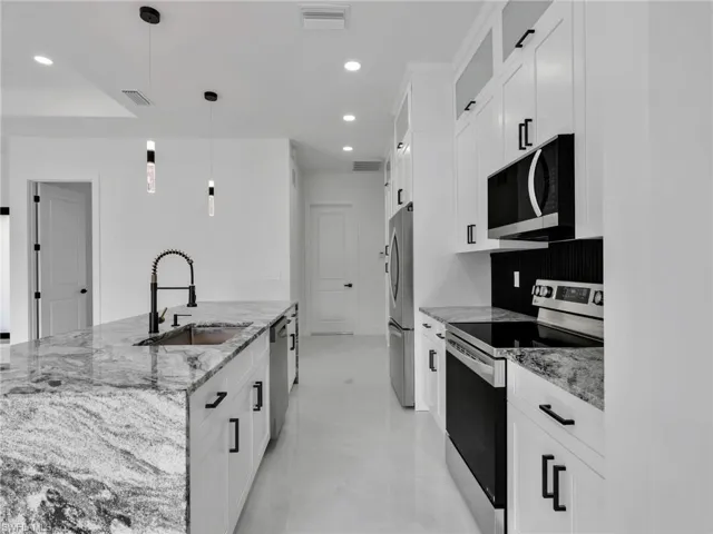 Kitchen featuring stainless steel appliances, light stone counters, a center island with sink, white cabinetry, and hanging light fixtures