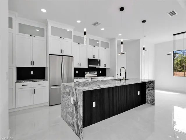 Kitchen with light stone counters, glass insert cabinets, a large island with sink, stainless steel appliances, and white cabinets