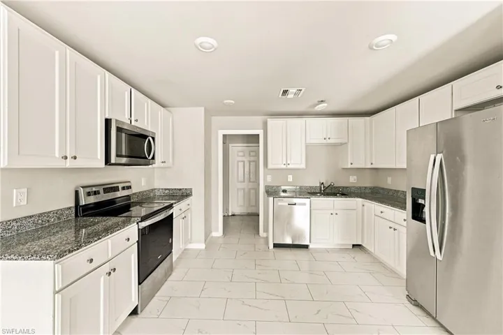 Galley-style kitchen featuring white cabinetry, stainless steel appliances including a side-by-side refrigerator and built-in microwave, granite countertops, tile flooring, and recessed ceiling lighting throughout