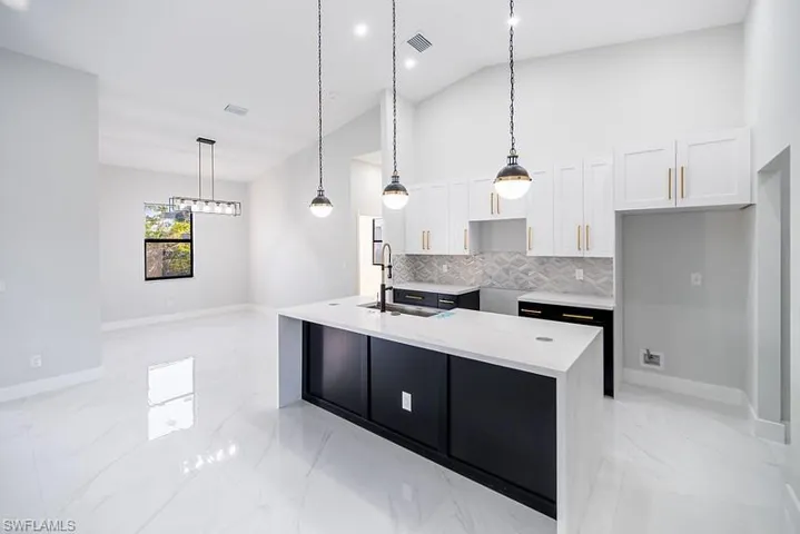 Dual tone kitchen with light stone countertops, decorative light fixtures, vaulted ceiling, a kitchen island with sink, and decorative backsplash