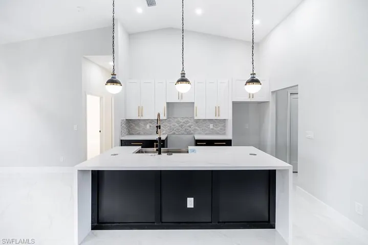 Kitchen featuring pendant lighting, light stone counters, a large island with sink, vaulted ceiling, and dark cabinetry