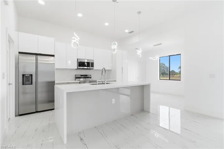 Kitchen with stainless steel appliances, white cabinetry, a kitchen island with sink, light stone counters, and decorative light fixtures