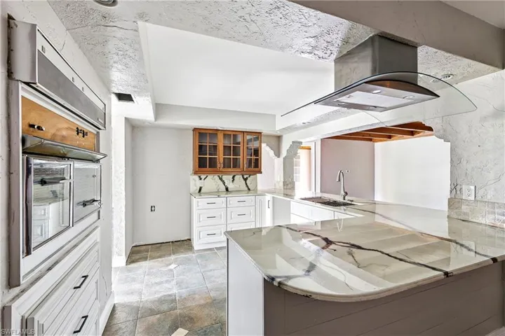 Kitchen featuring stone tile flooring, white cabinetry, a built-in oven, a range hood with a curved glass canopy, and a kitchen island with a light-toned countertop