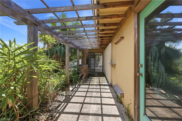 Exterior concrete walkway beneath a wooden pergola, bordered by lush tropical plantings
