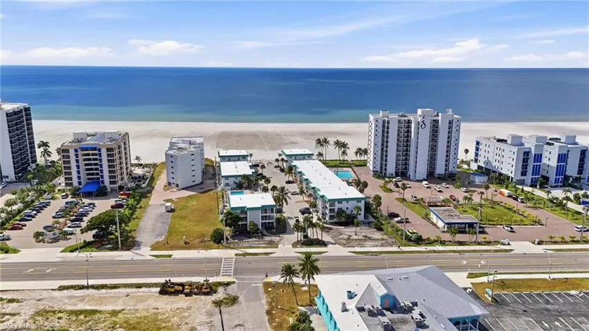 Coastal property featuring direct ocean frontage, expansive sandy beach, multi-story residential structures, paved parking areas, and tropical landscaping