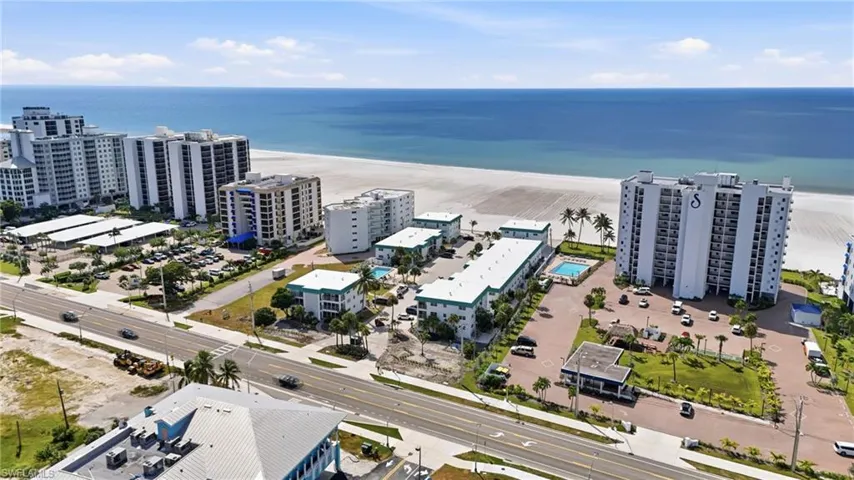 Aerial perspective of a multi-building condominium complex featuring direct shoreline access, multiple outdoor swimming pools, paved surface parking, and manicured grounds with palm trees