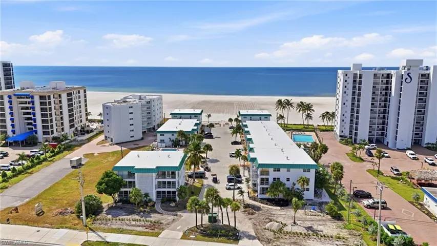 Aerial perspective of a multi-building residential complex featuring a swimming pool, paved surface parking, and mature palm trees with direct proximity to the ocean coastline
