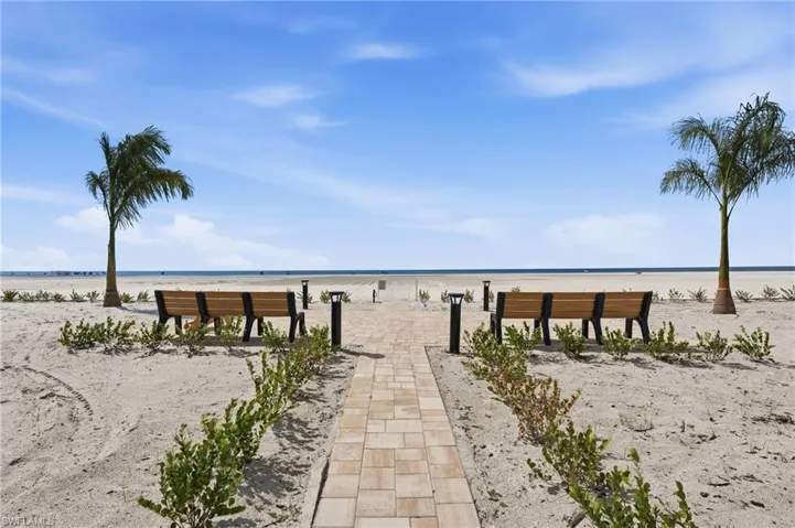 Paver walkway leading to sandy beachfront, featuring built-in wooden benches, low-profile landscape lighting, and palm trees