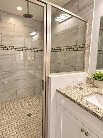 Walk-in shower featuring large-format wall tile, a mosaic tile floor, an integrated rainfall showerhead, and a glass enclosure, paired with a vanity featuring a speckled stone countertop and an undermount sink