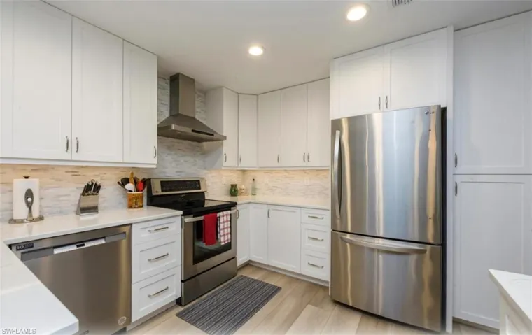 Kitchen with stainless steel appliances, wall chimney range hood, white cabinetry, backsplash, and light stone counters