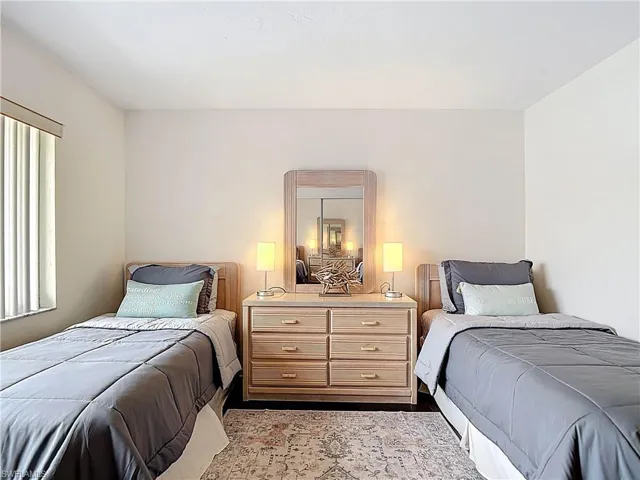 This bright room features neutral-toned walls, a window with vertical blinds, and dark flooring partially covered by an area rug