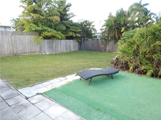 Rear yard featuring a grass lawn, square concrete pavers, perimeter wood fencing, and mature tropical landscaping