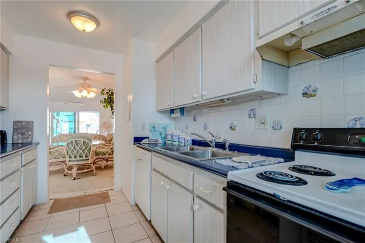 Kitchen with electric range oven, under cabinet range hood, decorative backsplash, light tile patterned flooring, and dark countertops
