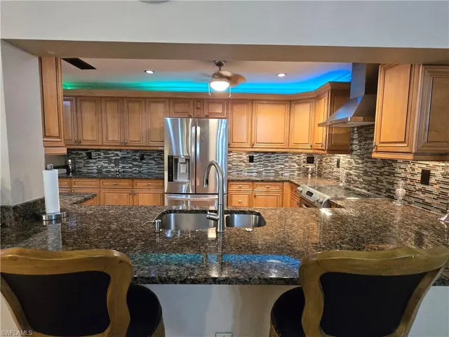 Kitchen with wall chimney range hood, stainless steel fridge with ice dispenser, ceiling fan, tasteful backsplash, and a breakfast bar area