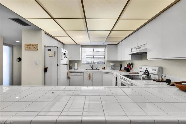Kitchen featuring tile counters, white appliances, under cabinet range hood, white cabinetry, and a drop ceiling