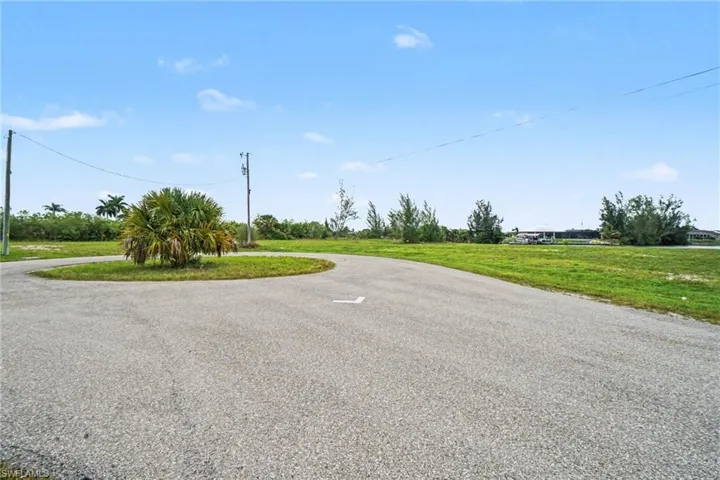 Expansive land parcel featuring a paved road, a circular median with a mature palm tree, and a clear blue sky
