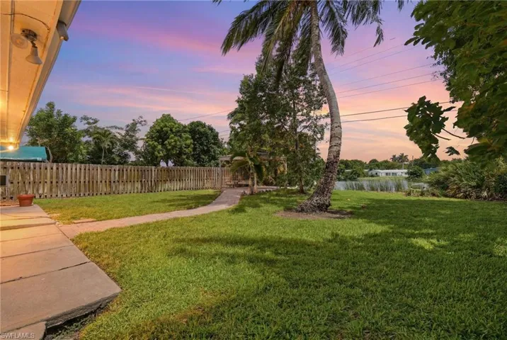 Expansive backyard featuring a manicured lawn, a winding paved pathway, a full privacy fence, and mature tropical landscaping