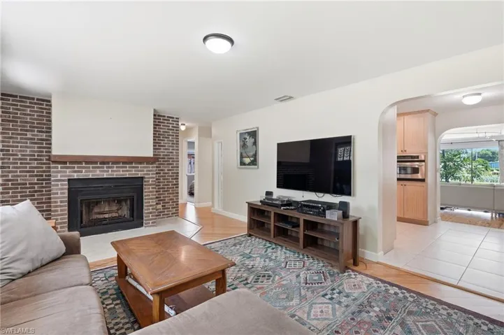 Living area featuring a brick fireplace with a wood mantle, light wood-finish flooring, and an arched doorway leading to a kitchen with light wood cabinetry and built-in appliances
