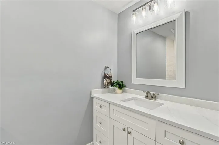 White vanity cabinetry featuring stone-composite countertop with integrated sink, polished metal faucet, rectangular framed mirror, and multi-bulb light fixture