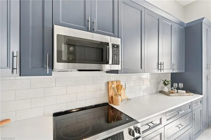 Shaker-style cabinetry in slate blue, white tile backsplash, light-toned solid surface countertops, and stainless steel built-in microwave