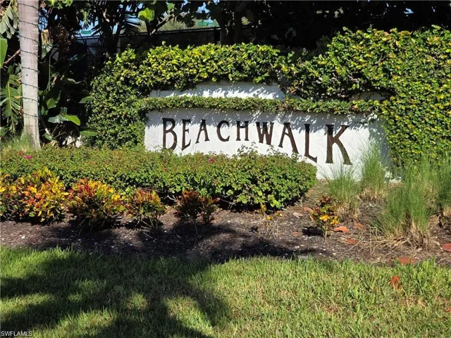 Stucco monument signage featuring metal lettering surrounded by mature hedges, palm trees, ornamental shrubbery, and landscaped mulch beds