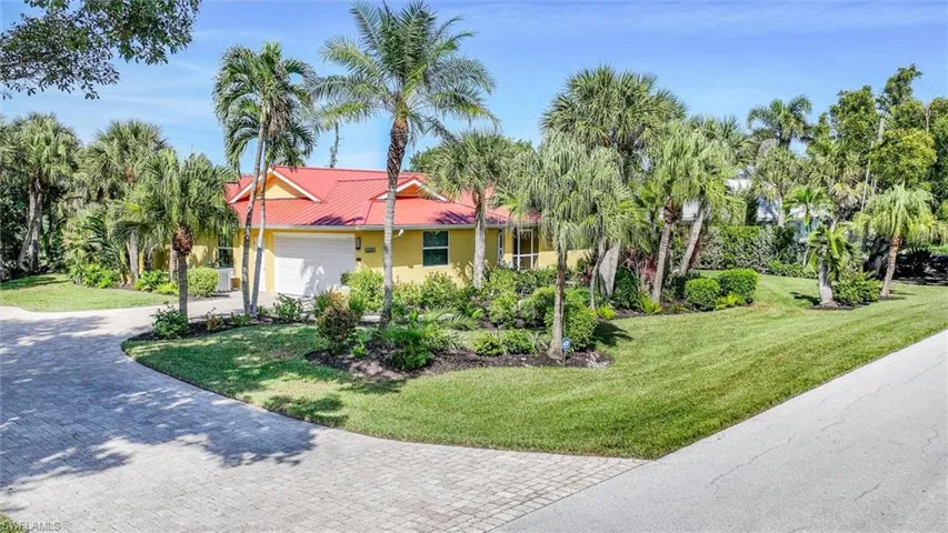Exterior of home featuring a red metal roof, attached two-car garage, and extensive landscaping with mature palm trees and paved driveway