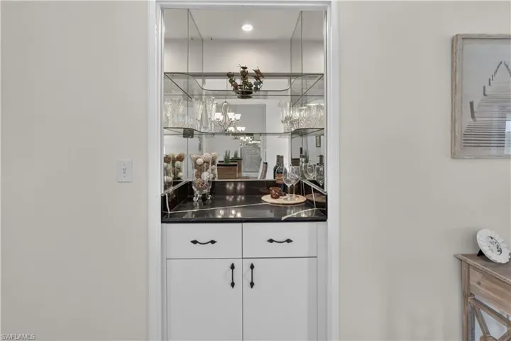Built-in bar with white cabinetry, black countertop, mirrored back wall, and glass shelving
