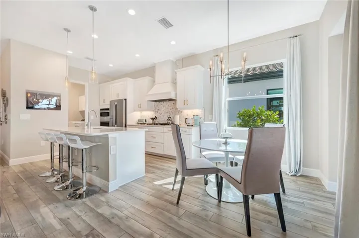 Kitchen with hanging light fixtures, custom range hood, light hardwood / wood-style flooring, an island with sink, and tasteful backsplash
