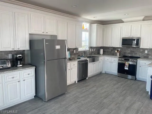 Kitchen featuring white cabinetry, stainless steel appliances, backsplash, ornamental molding, and light stone countertops