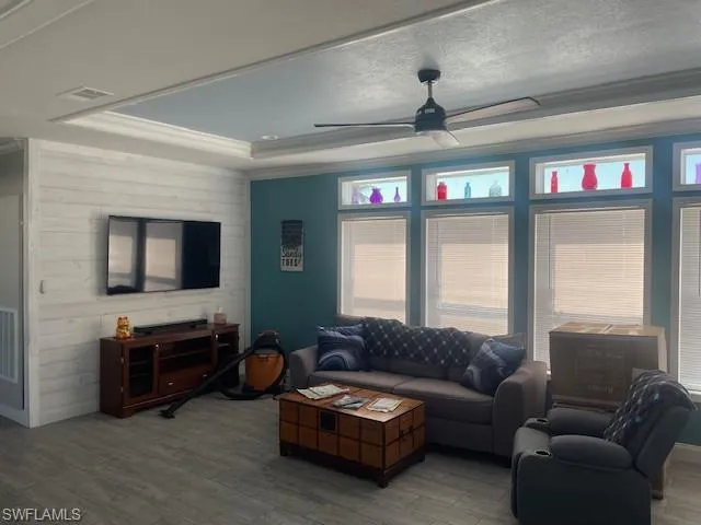 Living room with ceiling fan, wood finished floors, ornamental molding, a textured ceiling, and a tray ceiling