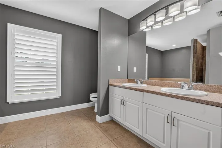 Bathroom featuring a double vanity with white cabinetry and an integrated sink, a large mirror, an overhead multi-bulb light fixture, a window with plantation shutters, and light-toned floor tiles