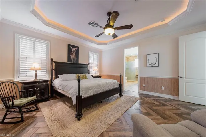 Bedroom featuring dark parquet flooring, ensuite bathroom, ceiling fan, ornamental molding, and a tray ceiling