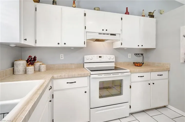 Kitchen with white electric stove, sink, light tile patterned flooring, and white cabinets