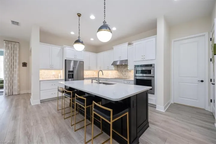 Upgraded Kitchen featuring custom lighting, quartz countertops, marble backsplash, white cabinetry, contrasting center island, cooktop, wall oven and under cabinet range hood
