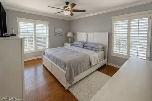 Hardwood-finish flooring featuring a white bed frame with a panel headboard, dual windows with plantation shutters, crown molding, and a ceiling fan with integrated lighting