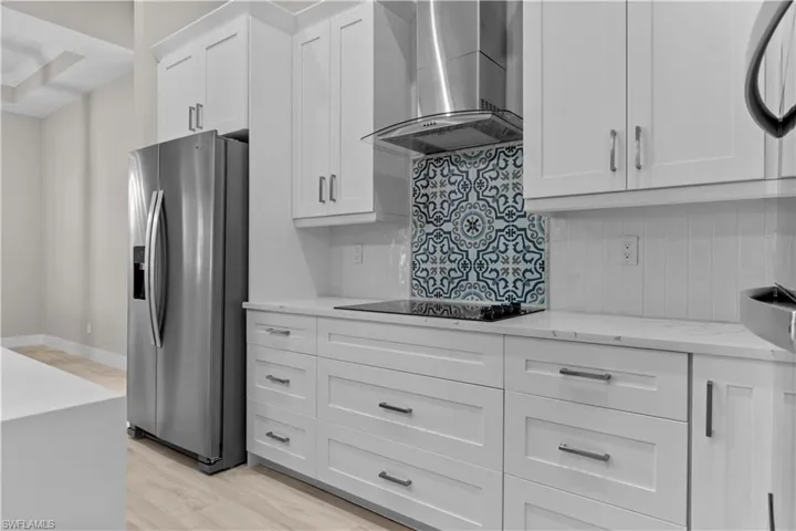 Kitchen with light wood-type flooring, wall chimney range hood, backsplash, light stone counters, and stainless steel fridge