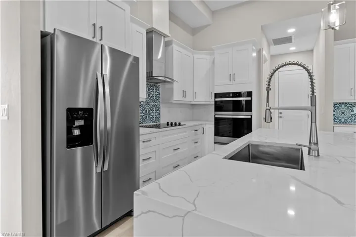 Kitchen featuring wall chimney range hood, backsplash, stainless steel appliances, light stone countertops, and decorative light fixtures