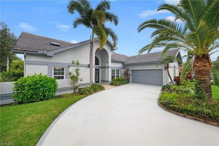Stucco exterior featuring an arched entryway, attached two-car garage, tiled roof, and paved driveway surrounded by mature palms and landscaping