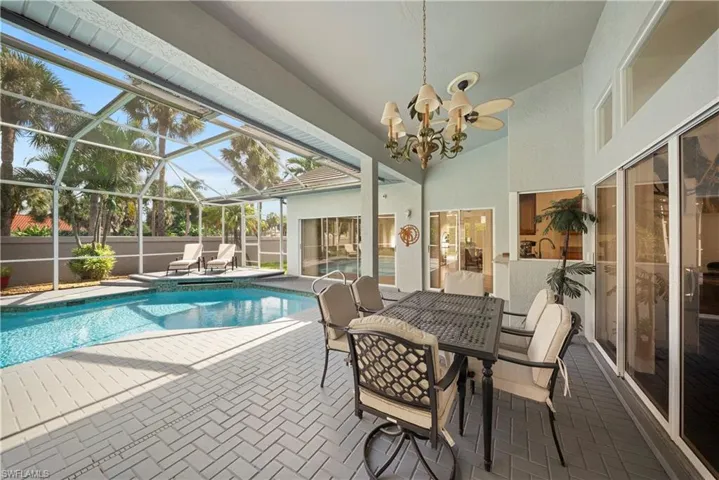 Covered patio with herringbone-patterned pavers, screened-in swimming pool enclosure, integrated pass-through window, and tiered chandelier with ceiling fan