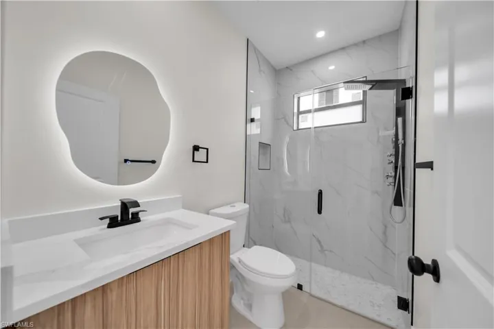 Modern bathroom featuring a custom-shaped backlit mirror, wood-finish vanity with a white countertop, and a glass-enclosed shower with large format tile surround