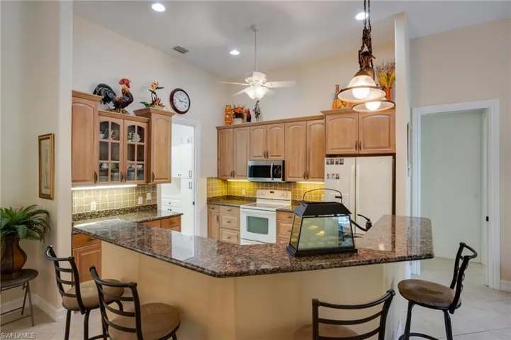 Kitchen featuring a breakfast bar area, white appliances, dark stone counters, decorative light fixtures, and a ceiling fan