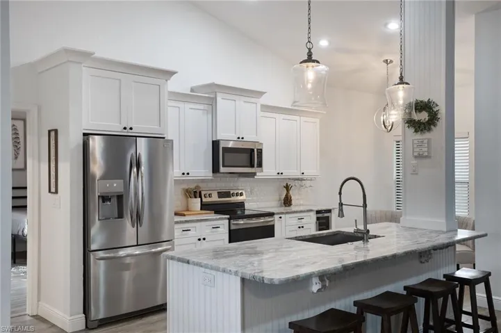 Kitchen featuring an island with sink, light stone counters, white cabinets, hanging light fixtures, and appliances with stainless steel finishes