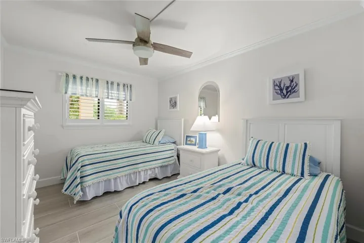 Bedroom featuring two single beds, wood-finish flooring, a ceiling fan, crown molding, and a window with plantation shutters