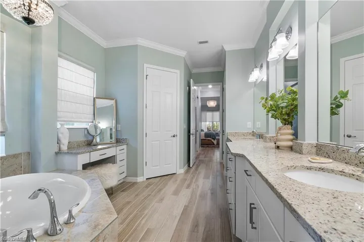 Dual vanities with stone countertops, wood-finish flooring, built-in soaking tub, and crown molding