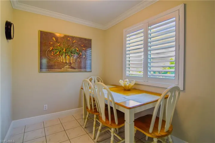 Dining room with impact glass window, plantation shutters, 5 1/4 baseboards, and crown moulding just outside of kitchen including white and wooden dining table and chairs