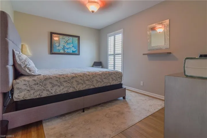 Primary bedroom with upholstered bed, warm wooden flooring, ceiling fan, impact glass window with plantation shutter