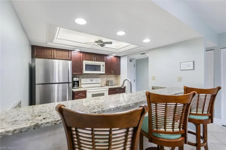Kitchen with white appliances, light stone countertops, a kitchen breakfast bar, backsplash, and a raised ceiling