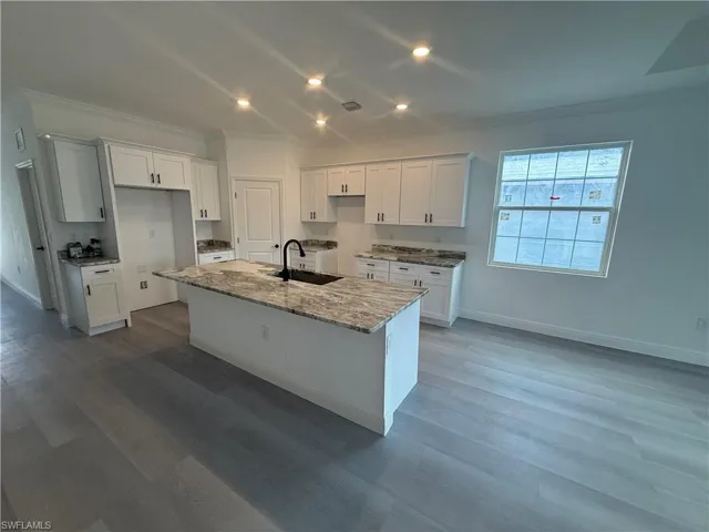 Kitchen with ornamental molding, a center island with sink, white cabinetry, light stone countertops, and recessed lighting
