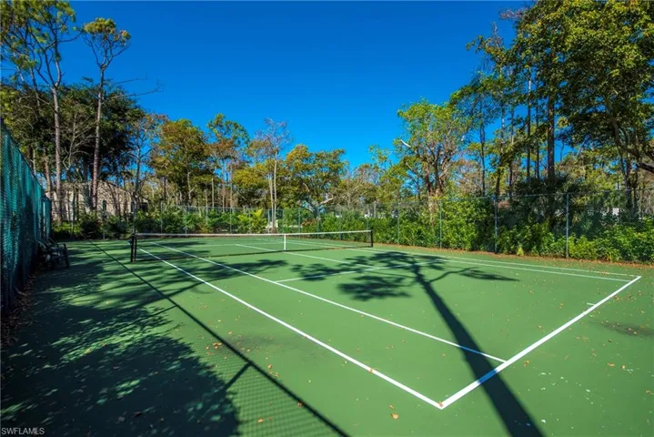 Private tennis court featuring a green surface with white line markings, surrounded by fencing and mature trees
